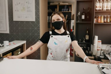 Tıbbi yüz maskesi takmış, dövmeleri olan mutlu bir bayan barista gülümseyip kahve dükkanında müşterilerini bekliyor. Kafenin güzel bir kadın sahibi barın arkasında kollarını açmış poz veriyor.. 