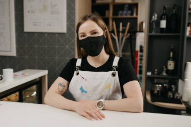Tıbbi yüz maskesi takmış, dövmeleri olan mutlu bir bayan barista gülümseyip kahve dükkanında müşterilerini bekliyor. Kafenin güzel bir bayan sahibi barın arkasında kollarını kavuşturmuş poz veriyor.. 