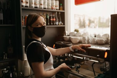 Siyah maskeli, dövmeli, mutlu bir bayan barista kafede bir fincan kahve yapar. Kahve makinesinin yanında poz veren bir garson.. 