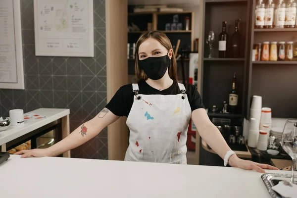 A happy female barista with tattoos in a medical black face mask smiles and waiting for clients in the coffee shop. A beautiful female owner of the cafe poses with her arms wide apart behind the bar.