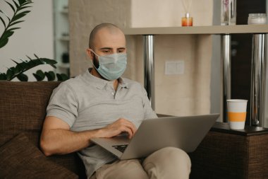 Uzaktan kumanda. Tıbbi maske takan bir adam karantina sırasında koronavirüsü yaymamak için uzaktan kumandayla çalışıyor. COVID-19 salgını sırasında evden çalışan sakallı bir adam..