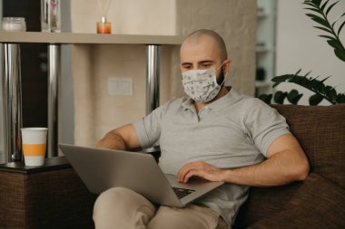 Uzaktan kumanda. Tıbbi maske takmış bir adam karantina sırasında koronavirüsten kaçmak için bilgisayarında uzaktan çalışıyor. COVID-19 salgını sırasında bir işadamı evden çalışır..