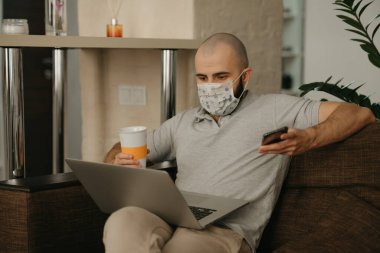 Yüzü maskeli bir adam karantina sırasında koronavirüsü yaymamak için uzaktan kumandayla çalışıyor. Bir adam COVID-19 salgını sırasında elinde telefon ve bir fincan çayla evde çalışıyor..