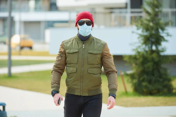 一个戴着医疗面罩的男人 为了避免在舒适的街上拿着智能手机而被传播开来 一个走路的人戴着红帽子 太阳镜 戴着防毒面具 — 图库照片
