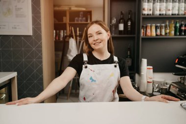 Barın arkasında beyaz önlüklü bir bayan barista bir kafede poz veriyor. Bir kahve dükkanında kolları açık duran ve gülümseyen güzel bir kadın işyeri sahibi..