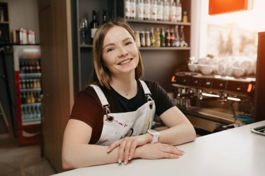 Beyaz önlüklü bayan bir barista bir kafede tezgaha yaslanmış poz veriyor. Kahve dükkanında kollarını kavuşturmuş gülümseyen ve ayakta duran güzel bir kadın işyeri sahibi..