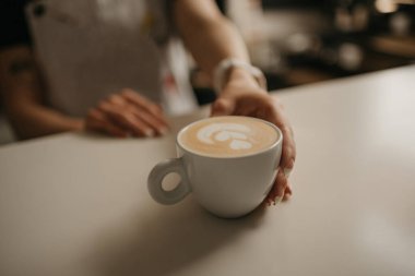 Bayan bir barista, bir kafede müşterisine bir fincan sıcak latte uzatıyor. Müşteri siparişi hazırlayan bir garson.