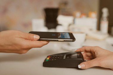 Bir kafede kontaksız Ödeme PASS teknolojisiyle latte almaya hazırlanan bir akıllı telefonun yakın plan fotoğrafı. Kadın bir barista, bir kahve dükkanında müşteriye ödeme yapmak için bir terminali soyar..