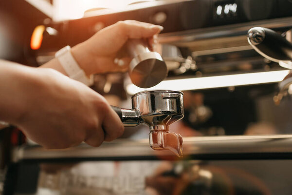 A close photo of female hands which holds a metal tamper and a portafilter with coffee in a coffee shop. A barista preparing for pressing ground coffee for brewing espresso or americano in a cafe.
