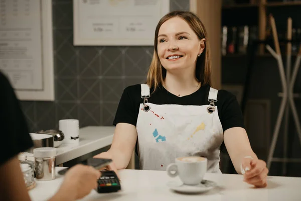 A close portrait of a female barista who smiling holds out to a client a terminal for paying. A woman paying for a cup of latte with a smartphone by contactless PAY PASS technology in a cafe.