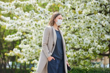 Koronavirüsten korunmak için tıbbi maske takan bir kız parkta akıllı telefon tutuyor. COVID-19 'a karşı maskeli bir kadın çiçek açan ağaçlar arasındaki sosyal mesafeyi koruyor..