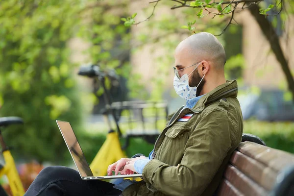 A man in a medical face mask to avoid the spread coronavirus works remotely on a laptop in the park. A guy wears a pilot sunglasses sits on a bench on the street with a computer near a bicycle.