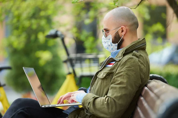 A man in a face mask to avoid the spread coronavirus works remotely on a laptop in the park. A guy wears sunglasses sits on a bench on the street with a computer near a bicycle. White-red-white flag.