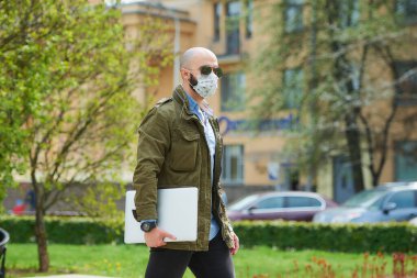 Tıbbi yüz maskesi takmış kel bir adam parkta dizüstü bilgisayarla dolaşmaktan kaçınıyor. Şehrin sokaklarında bir adam n95 yüz maskesi ve pilot güneş gözlüğü takıyor..
