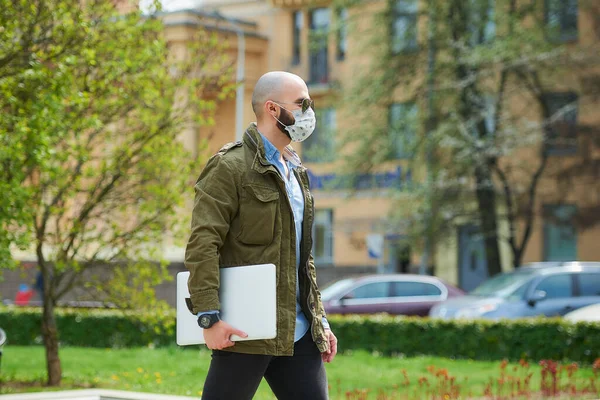 A bald man with a beard in a medical face mask to avoid the spread coronavirus walks with a laptop in the park. A guy wears n95 face mask and a pilot sunglasses on the street of the city.