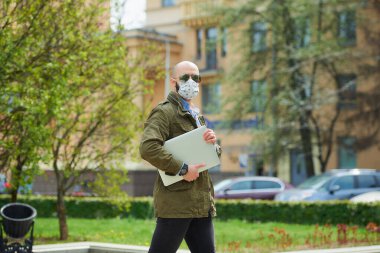 Tıbbi yüz maskesi takmış kel bir adam parkta dizüstü bilgisayarla dolaşmaktan kaçınıyor. Şehrin sokaklarında bir adam n95 yüz maskesi ve pilot güneş gözlüğü takıyor..