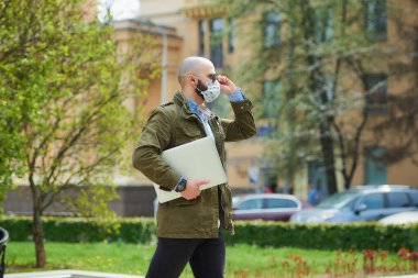Tıbbi yüz maskesi takmış kel bir adam parkta dizüstü bilgisayarla dolaşmaktan kaçınıyor. Şehrin sokaklarında bir adam n95 yüz maskesi ve pilot güneş gözlüğü takıyor..