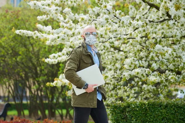 A bald man with a beard in a medical face mask to avoid the spread coronavirus walks with a laptop in the park. A guy wears n95 face mask and a pilot sunglasses in the garden between flowering trees.