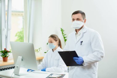 Ciddi bir cerrah tek kullanımlık eldiven ve solunum cihazı takar. Elinde hastane evrakları olan siyah bir dosya tutar. Kadın bir doktor köşedeki masaüstü bilgisayarının önünde oturuyor..  