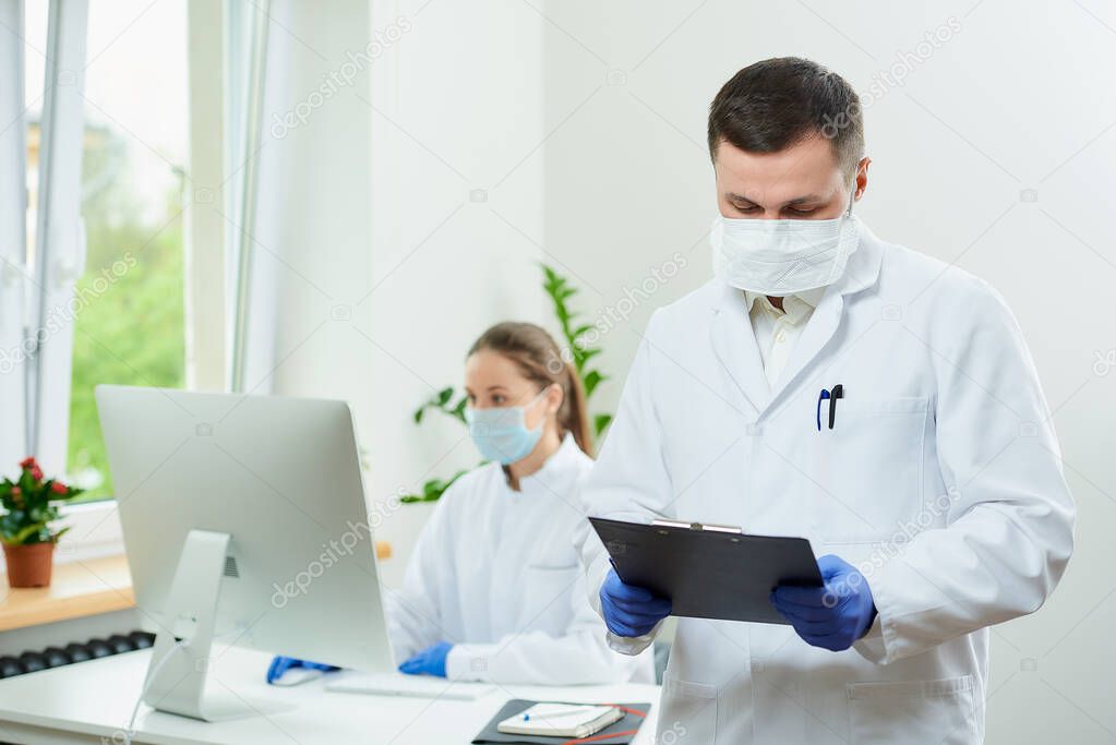 A surgeon with bristles wears a respirator holds a black clipboard and fills the patient's card in a hospital. A female doctor sitting in front of the desktop computer in the corner of the room 