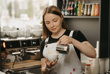 Elinde süt kupası olan kadın bir barista kafede kahve üzerine kahve sanatı yapıyor. Güzel bir barista, akşamları espressonun kremasını kırmamak için dar bir dereyle süt döküyor..