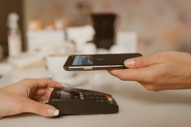 Bir kafede kontaksız Ödeme PASS teknolojisiyle latte almaya hazırlanan bir akıllı telefonun yakın plan fotoğrafı. Kadın bir barista, bir kahve dükkanında müşteriye ödeme yapmak için bir terminali soyar..