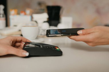 Bir kafedeki NFC teknolojisiyle akıllı telefonuyla kahvesini ödemeye hazırlanan bir kadın. Kadın bir barista, bir kahve dükkanında müşteriye ödeme yapmak için bir terminali soyar..