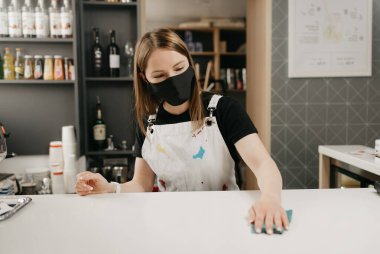 Siyah maskeli bir kadın barmen bir kahve dükkanında tezgahı ovuyor. Güzel bir garson kafeyi dezenfekte ediyor..