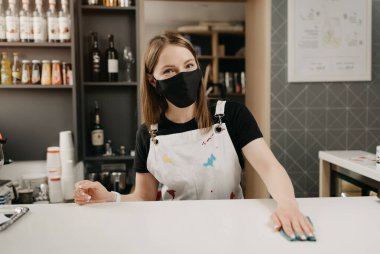 Tıbbi siyah maskeli bir bayan barista gülümsüyor ve bir kahve dükkanında tezgahı temizliyor. Güzel bir garson kafeyi dezenfekte ediyor..