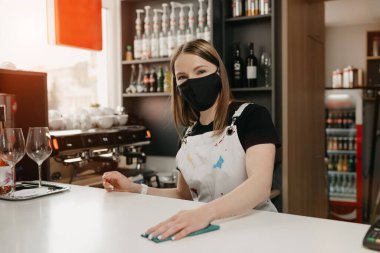 Tıbbi siyah maskeli bir bayan barista gülümsüyor ve bir kahve dükkanında tezgahı temizliyor. Güzel bir garson kafeyi dezenfekte ediyor..