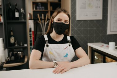Tıbbi siyah maskeli mutlu bir bayan barista gülümsüyor ve kahve dükkanında müşterilerini bekliyor. Kafenin güzel bir bayan sahibi barın arkasında kollarını kavuşturmuş poz veriyor.. 