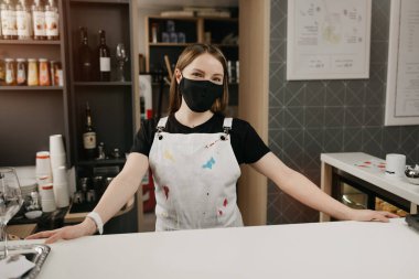 Tıbbi siyah maskeli mutlu bir bayan barista gülümsüyor ve kahve dükkanında müşterilerini bekliyor. Kafenin güzel bir kadın sahibi barın arkasında kollarını açmış poz veriyor..