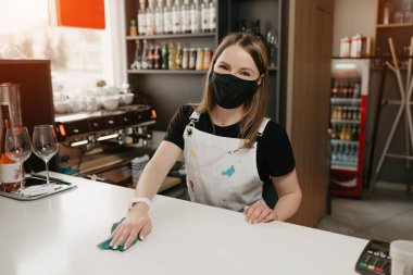 Siyah maskeli bir kadın barmen bir kahve dükkanında tezgahı ovuyor. Güzel bir garson kafeyi dezenfekte ediyor..