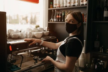Medikal siyah maskeli mutlu bir bayan barista kahvede kahve yapar ve gülümser.. 