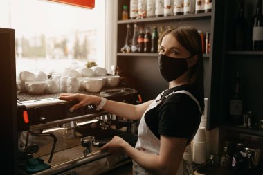 A happy female barista in a medical black face mask smiles and brews a cup of coffee in a coffee machine in the cafe.  clipart