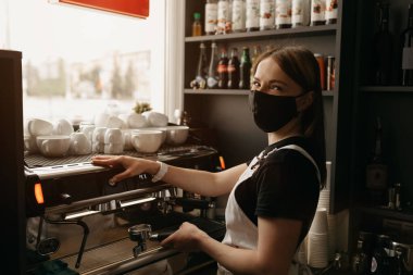 Tıbbi siyah maskeli mutlu bir bayan barista kafede bir fincan kahve yapar. Kahve makinesinin yanında poz veren bir garson.. 