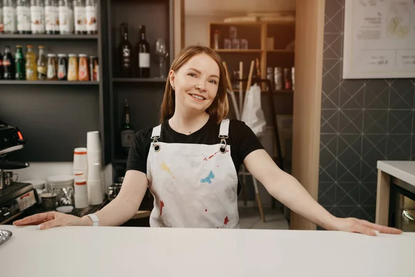 A female barista with a white apron poses behind the bar in a cafe. A pretty female business owner smiling and standing with her arms wide apart in a coffee shop.