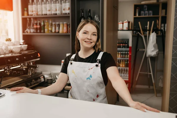 A female barista with a white apron poses behind the bar in a cafe. A pretty female business owner stands with her arms wide apart in a coffee shop.