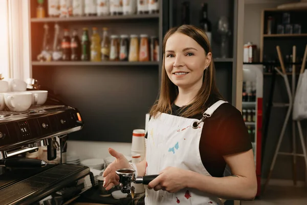 A female barista with white apron smiles and poses holding a metal tamper and a portafilter with coffee in a coffee shop. A pretty female business owner near a professional espresso machine in a cafe.