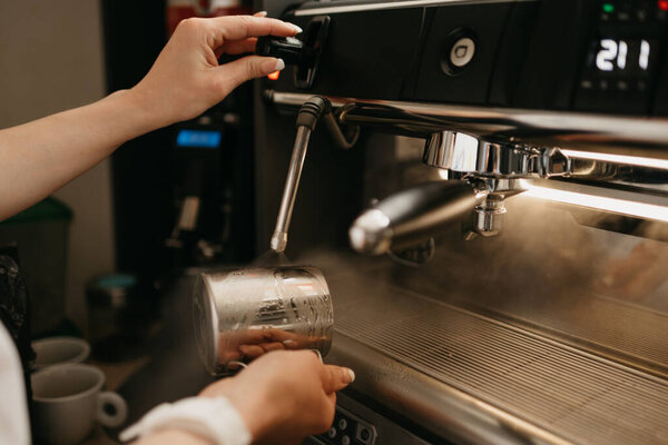 A female barista wears a white apron purges a metal mug with steam in a professional espresso machine in a coffee shop. A close-up photo of cleaning a cup in a coffee machine in a cafe.