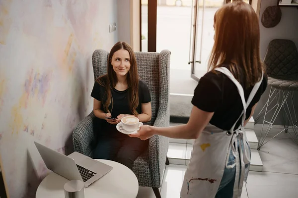 A female barista holds out a latte to a girl in a coffee shop. A woman with long hair working remotely on a laptop keeps social distance grabs a cup of coffee in a cafe.
