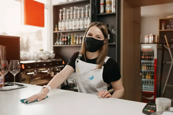 Tıbbi siyah maskeli bir bayan barista gülümsüyor ve bir kahve dükkanında tezgahı temizliyor. Güzel bir garson müşterileri beklerken bir kafeyi dezenfekte ediyor..
