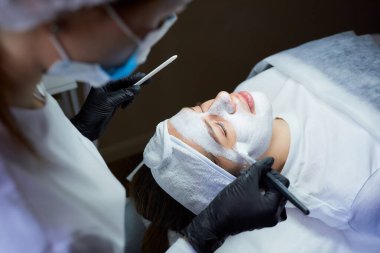 Kızların yüzüne gommage 'i soyan bir kozmetolog bakış açısı. Tıbbi maske takan ve tek kullanımlık siyah eldivenli bir doktor. Güzellik salonunda cilt temizleme ameliyatı sırasında.