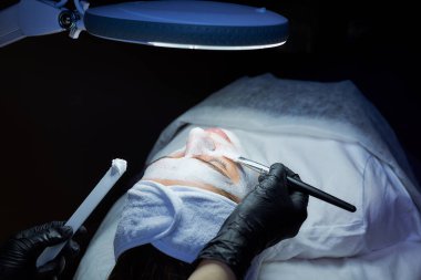 Hastanın kafasının tepesinden, kadının yüzünde fırçayla soyulan gommage 'in fotoğrafı. Güzellik salonunda cilt temizleme işlemi sırasında kozmetolojistin elleri..