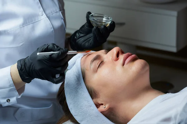 A female cosmetologist wearing black disposable medical gloves uses a brush to apply a superficial transparent face peeling to a girl. A cosmetology procedure in a beauty salon for skin cleaning.