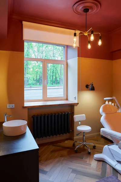 Interior of the cosmetology office with an electrical facial beauty bed and chair, a salon stool, a trolley cart with skincare products, a led lamp and a sink in beauty salon. A window curtain raised.