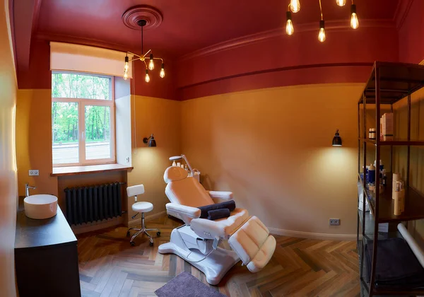 Wide photo of an interior of the cosmetology office with a sink, a full-electrical facial beauty bed and chair, a salon stool, a trolley cart with skincare products, a led lamp, and metal shelves.