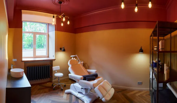 Interior of the cosmetology office with a sink, a full-electrical facial beauty bed and chair, a salon stool, a trolley cart with skincare products, and a led lamp in a salon. A window curtain raised.