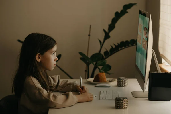 Distance learning. A young girl with long hair studying remotely online. A female child learns a lesson using an all-in-one computer at home. Home education.