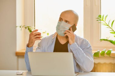 Tıbbi maske takmış bir adam karantina sırasında koronavirüsten kaçmak için bilgisayarında uzaktan çalışıyor. Kel bir adam COVID-19 salgını sırasında evden ararken elinde kahve tutuyordu..